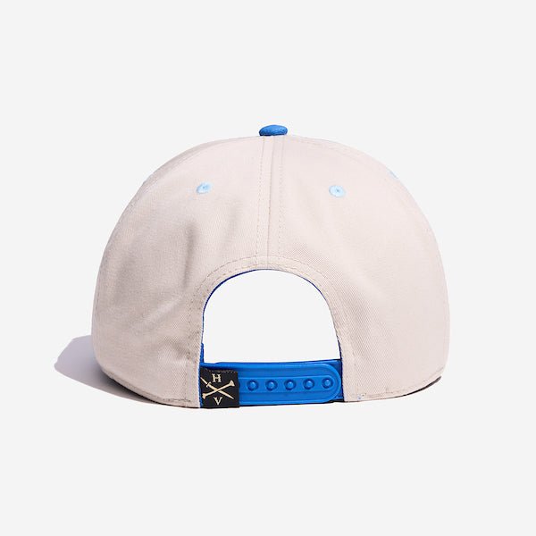 The back of a blue and white hat with a blue adjustable strap. Next to the strap is a small square fabric tag that displays the letters H and V.