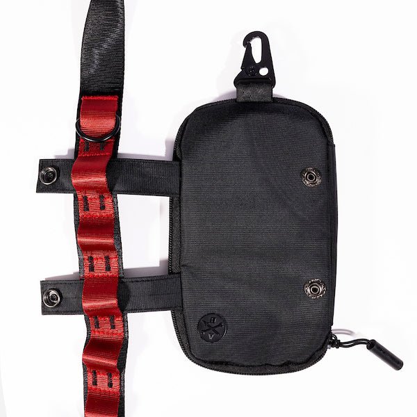 The photo shows a close up detailing of the shoulder strap. Featured is the Blue decorative detailing. The detailing has loops for attaching clips and a tech case. The tech case is pictured to the right side displaying the straps going through the loops. The top loop has a "D"-Ring attached.