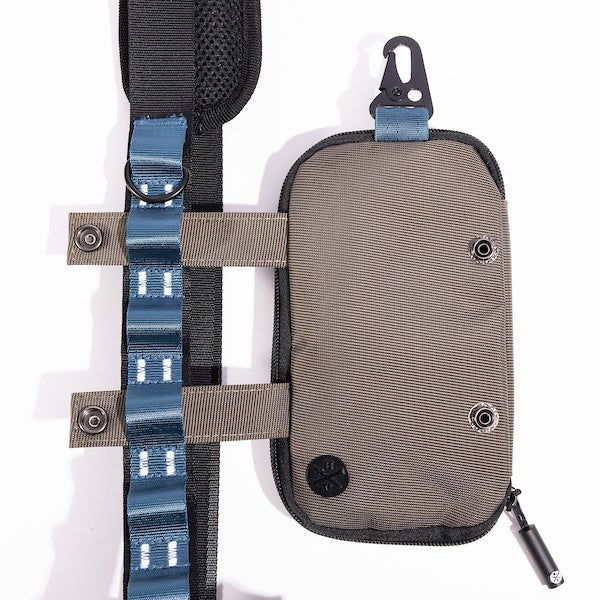 The photo shows a close up detailing of a shoulder strap. Featured is the Blue decorative detailing. The detailing has loops for attaching clips and a tech case. The tech case is pictured to the right side displaying the straps going through the loops. The top loop has a "D"-Ring attached.