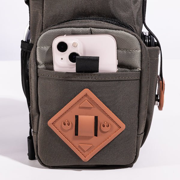 The photo shows a close up of the bags main outer zipper pouch. The pouch has an orange diamond shaped rubber patch on the front. There is a pocket on this pouch showing an iphone being held.