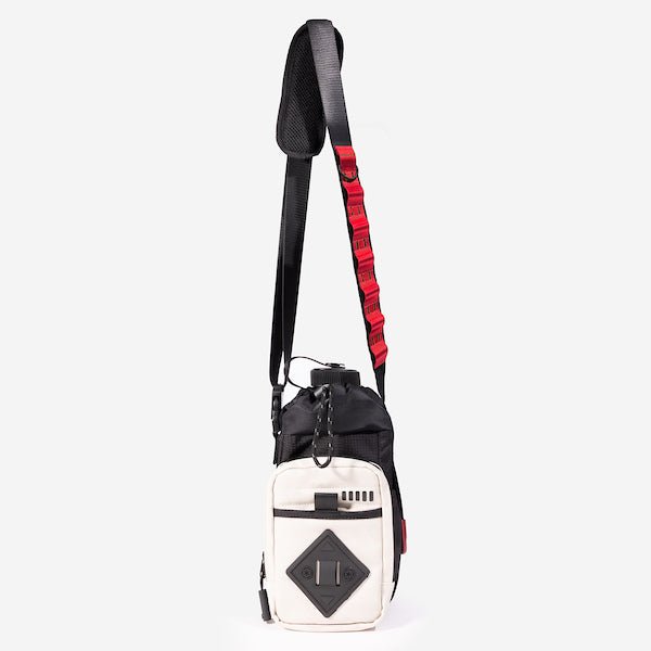 The photo shows a crossbody bag designed for carrying a water bottle. The bag features a drawstring closure at the top, a front zippered pocket, and a black rubber patch with a geometric design meant for attaching a clip. The adjustable black strap has a padded section for comfort and a red cord with holes to attach clips and a tech organizer bag (not pictured.) The bag is black with a front white zipper pouch and red detailing.