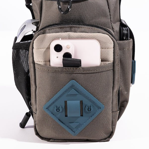 The photo shows a close up of the bags main outer zipper pouch. The pouch has a blue diamond shaped rubber patch on the front. There is a pocket on this pouch showing an iphone being held.