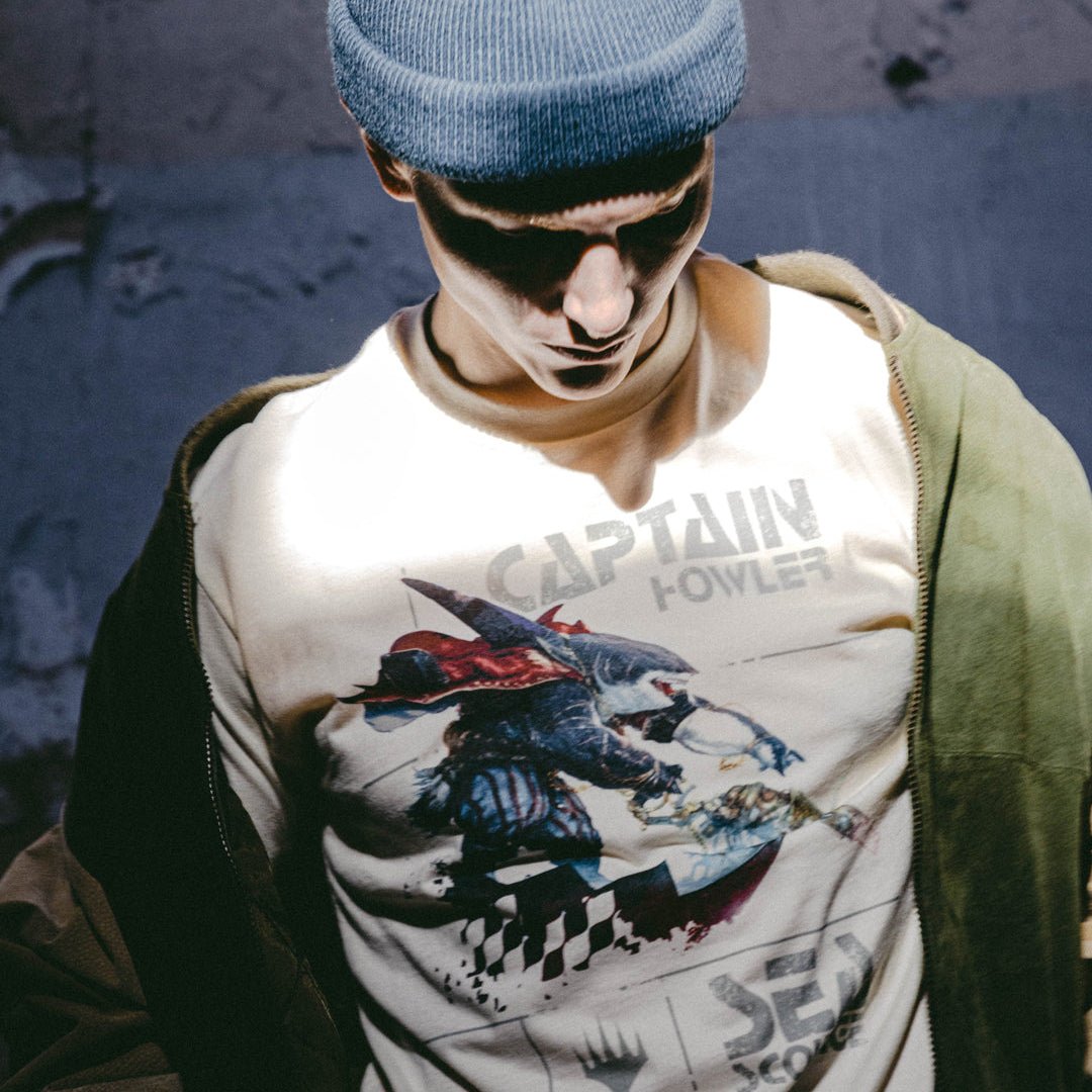 A male model in a beanie and hoodie with a grey background the main focus is the natural Magic: The Gathering t-shirt featuring Captain Howler, a fierce shark-like pirate with armored plating, a flowing red cape, and striped pants. He wields an ornate, steampunk-inspired harpoon gun, chained to his weapon. The background includes a distressed, grunge-style design with the words 'Captain Howler' and 'Sea Scourge' in bold, stylized font, along with the Magic: The Gathering Planeswalker symbol.