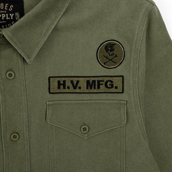 The photo shows a close up detail of a green shirt jacket. The Photo is featuring two patches above a shirt pocket. The top patch is circular and features a skull and cross bones logo. The bottom patch has the text "H.V. MFG."