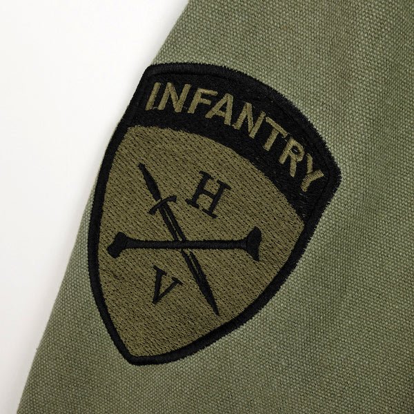 The photo shows a close up of a patch on the sleeve of a jacket. The patch is shield shaped and features the word "infantry" on the top. Below the word infantry is the Heroes and Villains Logo. The logo features a Diagonal sword overlapped with a diagonal femur bone in the opposite direction. The Letter "H" is above the sword and bone and the "V" is below the sword and bone.