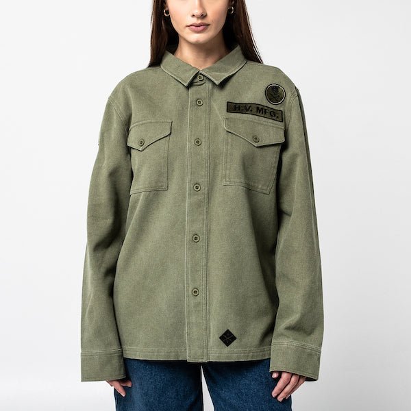 The photo shows a woman in a military style shirt jacket. The jacket is a thick canvas material and features 2 patches on the right chest. The jacket has buttons as a closer and 2 chest pockets.
