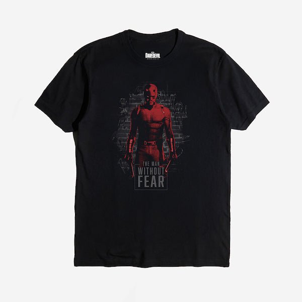 The photo is of a black tshirt. The tee features a stylized illustration of Daredevil, the Marvel Comics superhero. Daredevil is standing against a dark, graffiti-covered brick wall. His red suit is highlighted in a gritty, high-contrast design with shadows obscuring parts of his face. He holds his signature billy clubs in each hand, exuding a strong and fearless stance. The words 'THE MAN WITHOUT FEAR' are prominently displayed at the bottom
