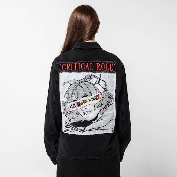 Woman standing backwards to show the back a black denim jacket. The jacket displays a large embroidered patch featuring an anime-style character with goggles labeled "The Mighty Nein." The character has spiky hair and a confident smile, with the words "CRITICAL ROLE" embroidered in red above the patch.