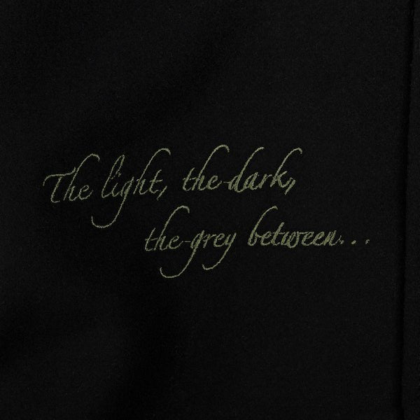 The photo shows up close of green embroidered text on black fabric. It says "The Light, The Dark, The Grey Between"