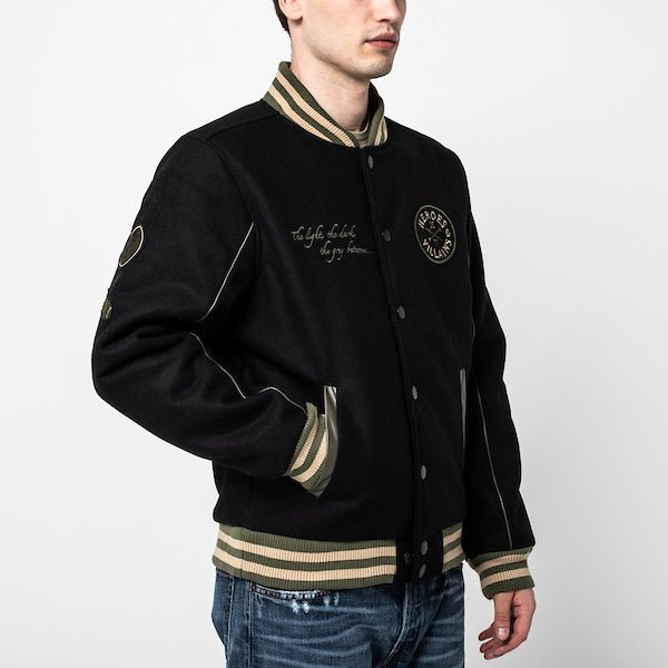 The photo shows a man facing right in a black jacket. The jacket features a green & beige striped cuff on the neckline, the wrists and the hem. The Jacket is a snap button closure. There is 2 pockets, embroidered text on the left chest and a patch on the right patch.