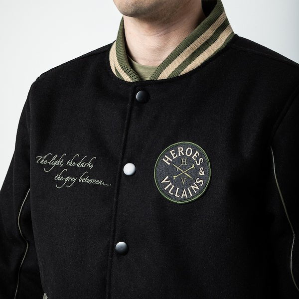 The photo shows a man in a black jacket. The jacket features a green & beige striped cuff on the neckline. The Jacket is a snap button closure. There is embroidered text on the left chest and a patch on the right patch.
