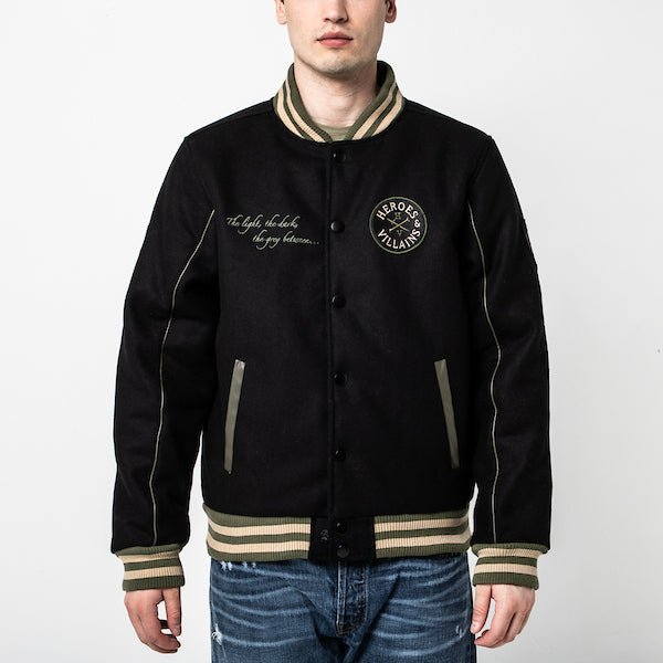 The photo shows a man in a black varsity jacket. The jacket features a green & beige striped cuff on the neckline, the wrists and the hem. The Jacket is a snap button closure. There is 2 pockets, embroidered text on the left chest and a patch on the right patch.