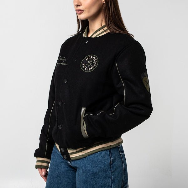 The photo shows a woman facing left in a black jacket. The jacket features a green & beige striped cuff on the neckline, the wrists and the hem. The Jacket is a snap button closure. There is 2 pockets, embroidered text on the left chest and a patch on the right patch.