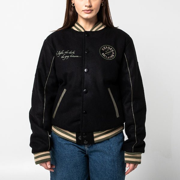 The photo shows a woman in a black varsity jacket. The jacket features a green & beige striped cuff on the neckline, the wrists and the hem. The Jacket is a snap button closure. There is 2 pockets, embroidered text on the left chest and a patch on the right patch.