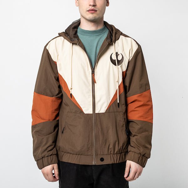 A man in a Star Wars-themed hooded windbreaker jacket featuring a Rebel Alliance emblem on the left chest. The jacket has a color-blocked design in brown, beige, and orange, with a front zipper, adjustable drawstrings on the hood, elastic cuffs, and two zippered pockets. The overall aesthetic is inspired by the Rebel pilot uniforms from the Star Wars universe