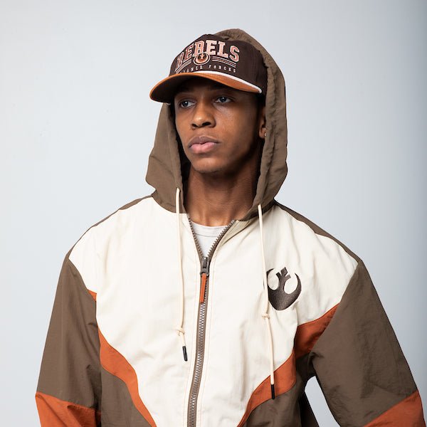 A man in a Star Wars-themed hooded windbreaker jacket and hat. The windbreaker features a Rebel Alliance emblem on the left chest. The jacket has a color-blocked design in brown, beige, and orange, with a front zipper, adjustable drawstrings on the hood, elastic cuffs, and two zippered pockets. The overall aesthetic is inspired by the Rebel pilot uniforms from the Star Wars universe