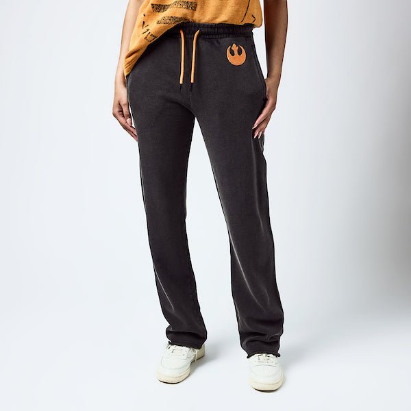 Woman in dark grey sweatpants with an elastic waistband and orange drawstring, featuring a small orange rebel logo near the left pocket and a vertical "rebel" graphic printed in distressed black text along the lower left leg.