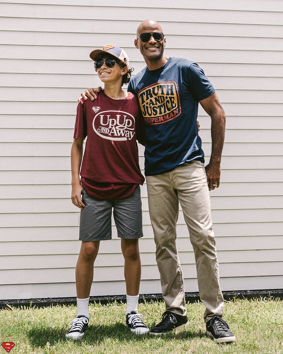 Father and Son standing outside of house. The son is wearing a superman hat and a red superman shirt that says "up up and away" The Dad is wearing a navy superman shirt that says "Truth and Justice"