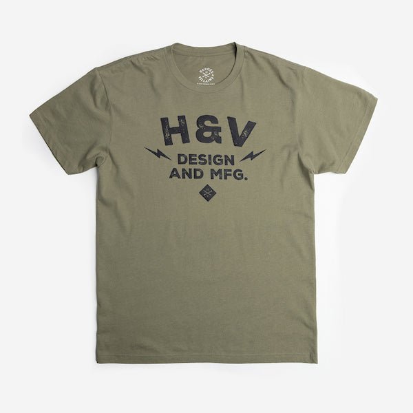 The photo shows a green short sleeve shirt. The print on the shirt says "H&V Design and MFG." The text is stacked with "H&V" on top, "Design" in the middle and "And MFG." below. On either side of the word "design" is a small lightening bolt shape. Under the text "And MFG" is a small diamond shape with a cross bones logo.