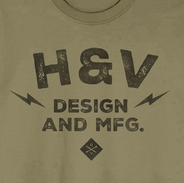 The photo shows a close up of a shirts print. The print on the shirt says "H&V Design and MFG." The text is stacked with "H&V" on top, "Design" in the middle and "And MFG." below. On either side of the word "design" is a small lightening bolt shape. Under the text "And MFG" is a small diamond shape with a cross bones logo.