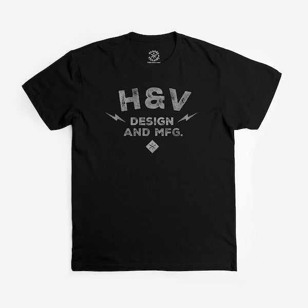 The photo shows a black short sleeve shirt. The print on the shirt says "H&V Design and MFG." The text is stacked with "H&V" on top, "Design" in the middle and "And MFG." below. On either side of the word "design" is a small lightening bolt shape. Under the text "And MFG" is a small diamond shape with a cross bones logo.