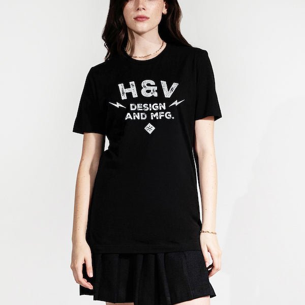 The photo shows a woman in a black short sleeve shirt. The print on the shirt says "H&V Design and MFG." The text is stacked with "H&V" on top, "Design" in the middle and "And MFG." below. On either side of the word "design" is a small lightening bolt shape. Under the text "And MFG" is a small diamond shape with a cross bones logo.