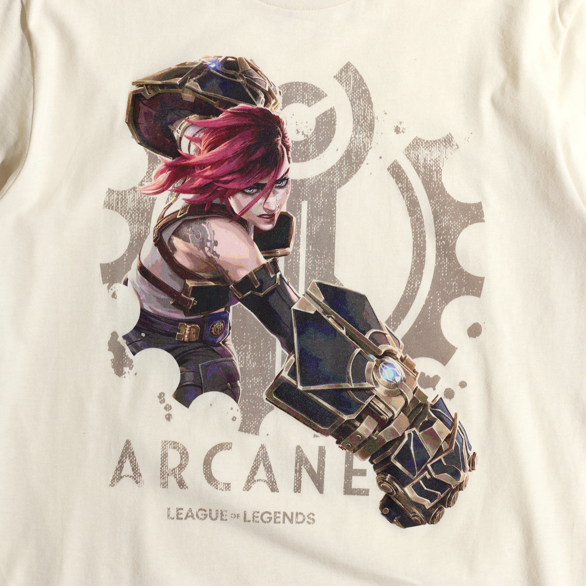 A cream-colored Arcane x League of Legends t-shirt featuring Vi in an action pose. She has her signature pink hair and wields her massive Hextech gauntlets, detailed with metallic and glowing blue accents. Behind her, a faded Piltover gear emblem adds depth to the design. 'Arcane' and 'League of Legends' are printed below in a bold, distressed font