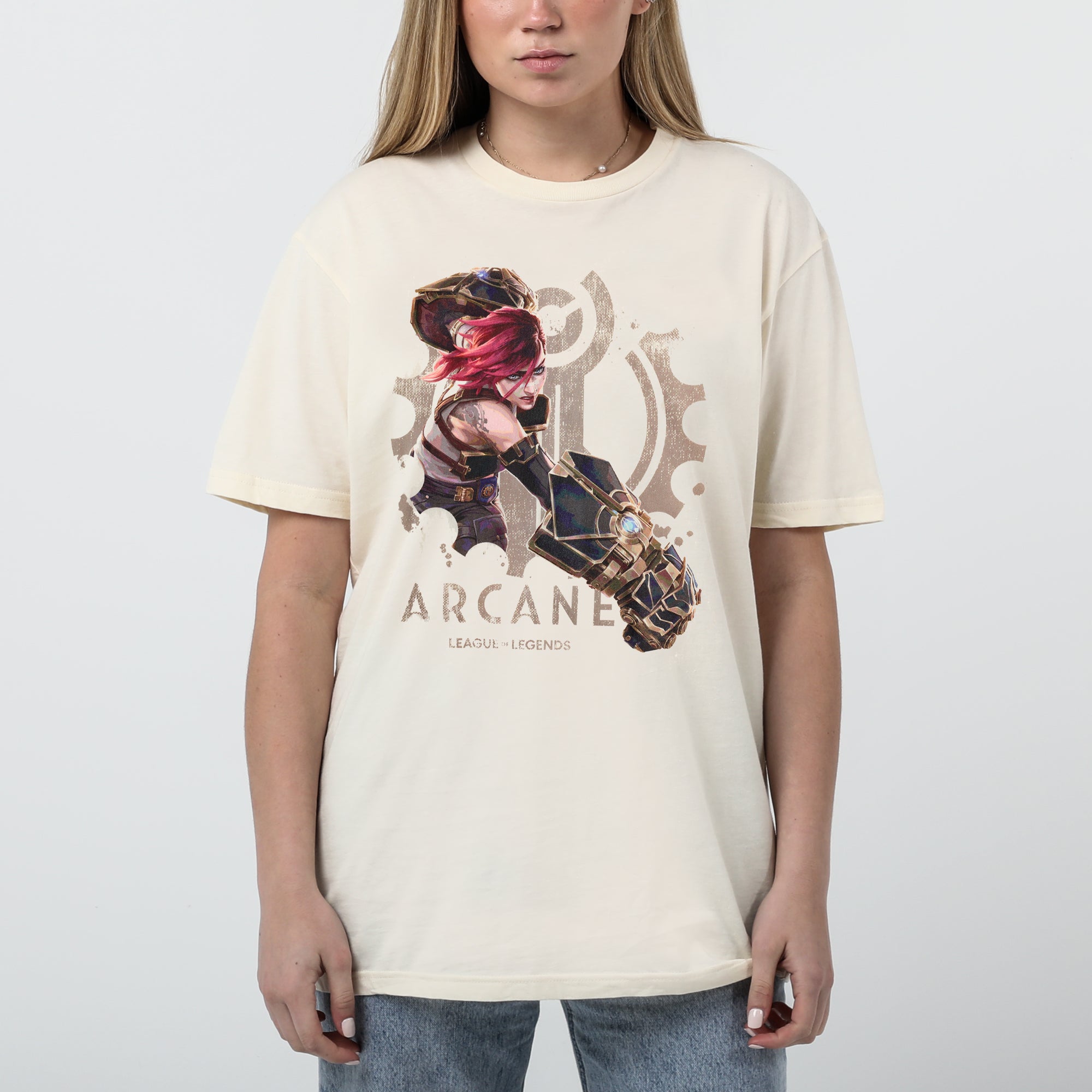A female model wearing a cream-colored Arcane x League of Legends t-shirt featuring Vi in an action pose. She has her signature pink hair and wields her massive Hextech gauntlets, detailed with metallic and glowing blue accents. Behind her, a faded Piltover gear emblem adds depth to the design. 'Arcane' and 'League of Legends' are printed below in a bold, distressed font
