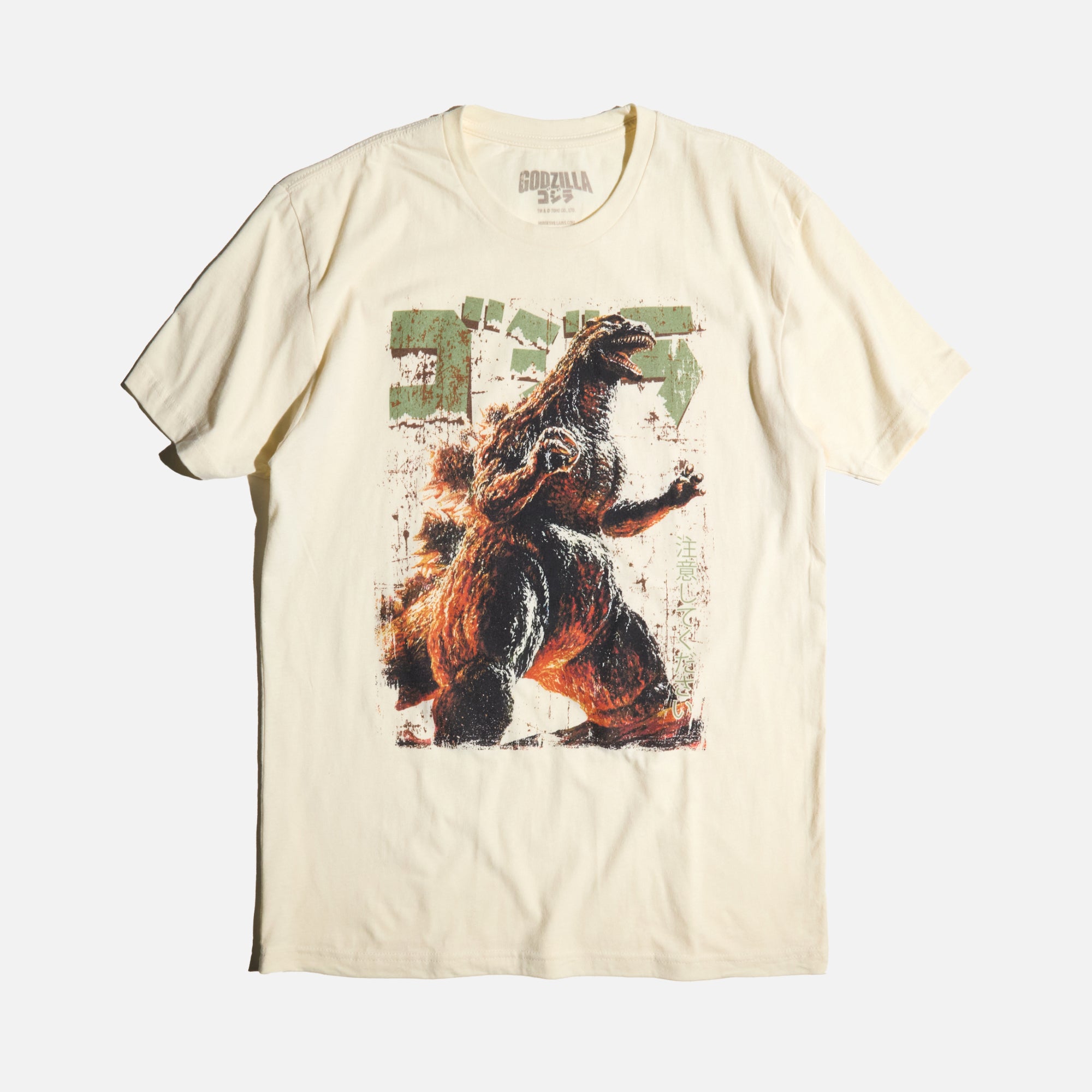 Natural-colored T-shirt featuring a vintage-style graphic of Godzilla roaring with a distressed design, Japanese text, and green block lettering.