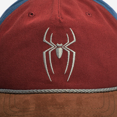 Close-up of a red snapback cap featuring a white embroidered spider logo on the front, with a brown suede-look brim and a blue fabric section visible at the top edge.
