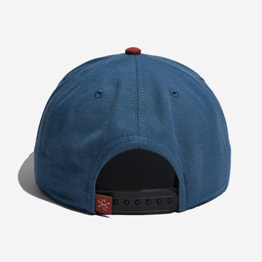 The back view of a blue snapback cap with a red button on top, a black adjustable strap, and a small red brand tag on the side.
