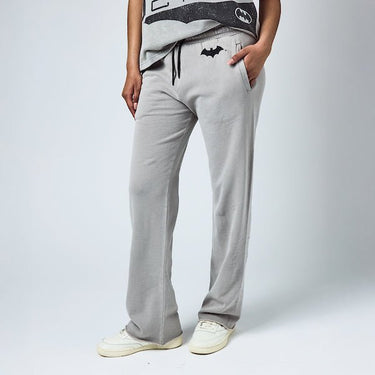 Woman in light gray sweatpants with an elastic waistband and black drawstring, featuring a small black Batman logo near the left pocket and a vertical "Gotham City" graphic printed in distressed black text along the lower left leg.