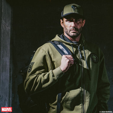 Man wearing a green The Punisher Zip-up hoodie and matching The Punisher Trucker Hat with a backpack, standing against a dark background.