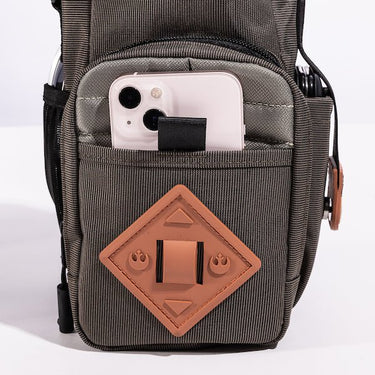 The photo shows a close up of the bags main outer zipper pouch. The pouch has an orange diamond shaped rubber patch on the front. There is a pocket on this pouch showing an iphone being held. 