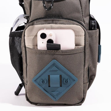 The photo shows a close up of the bags main outer zipper pouch. The pouch has a blue diamond shaped rubber patch on the front. There is a pocket on this pouch showing an iphone being held. 