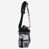 The photo shows a crossbody bag designed for carrying a water bottle. The bag features a drawstring closure at the top, a front zippered pocket, and a black molle loop strap meant for attaching a clip. The adjustable black strap has a padded section for comfort and a red cord with holes to attach clips and a tech organizer bag (Not pictured). The bag is in a subtle grey with monocromatic grey scale accents and pops of red. 