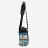 The photo shows a crossbody bag designed for carrying a water bottle. The bag features a drawstring closure at the top, a front zippered pocket, and a gold molle loop strap meant for attaching a clip. The adjustable black strap has a padded section for comfort and a gold cord with holes to attach clips and a tech organizer bag (Not pictured). The bag is in a subtle blue with blue monochromatic accents and pops of gold