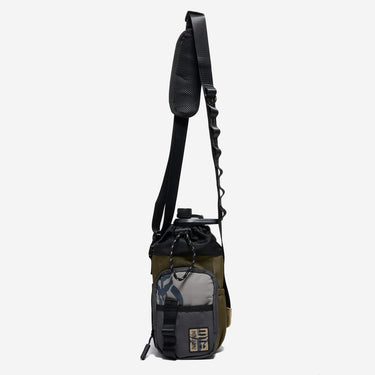 The photo shows a crossbody bag designed for carrying a water bottle. The bag features a drawstring closure at the top, a front zippered pocket, and black molle loop strap meant for attaching a clip. The adjustable black strap has a padded section for comfort and a brown molle loop strap to attach clips and a tech organizer bag (Not pictured). The bag is in a subtle brown with pops of grey and beige