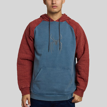 A man wearing a Spider-Man themed pullover hoodie with a blue body, red quilted sleeves, and a gray spider logo on the chest, set against a solid gray background.