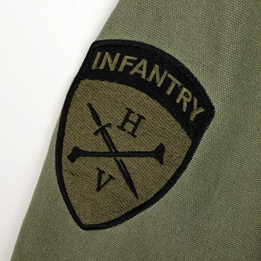 The photo shows a close up of a patch on the sleeve of a jacket. The patch is shield shaped and features the word "infantry" on the top. Below the word infantry is the Heroes and Villains Logo. The logo features a Diagonal sword overlapped with a diagonal femur bone in the opposite direction. The Letter "H" is above the sword and bone and the "V" is below the sword and bone. 