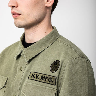 The photo shows a close up of a man facing the left. The photo is showing a close up detail of a green shirt jacket. The Photo is featuring two patches above a shirt pocket. The top patch is circular and features a skull and cross bones logo. The bottom patch has the text "H.V. MFG."