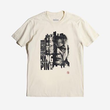 The photo is of an off-white colored tee. The tee features a gritty, high-contrast portrait of Wilson Fisk, also known as the Kingpin, with half of his face merging into a cityscape. His name, "Wilson Fisk," is stacked vertically, while "KINGPIN" and "OF CRIME" are displayed in bold, blocky typography. The design emphasizes his dominance over the criminal underworld, with the Daredevil "DD" logo in the lower corner.