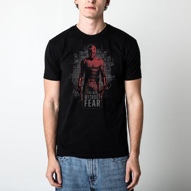 Man standing in a black tee. The tee features a stylized illustration of Daredevil, the Marvel Comics superhero. Daredevil is standing against a dark, graffiti-covered brick wall. His red suit is highlighted in a gritty, high-contrast design with shadows obscuring parts of his face. He holds his signature billy clubs in each hand, exuding a strong and fearless stance. The words 'THE MAN WITHOUT FEAR' are prominently displayed at the bottom