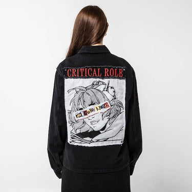 Woman standing backwards to show the back a black denim jacket. The jacket displays a large embroidered patch featuring an anime-style character with goggles labeled "The Mighty Nein." The character has spiky hair and a confident smile, with the words "CRITICAL ROLE" embroidered in red above the patch.
