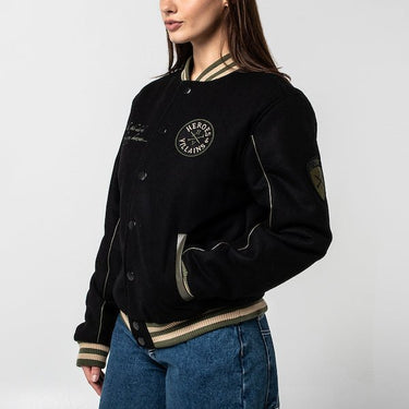 The photo shows a woman facing left in a black jacket. The jacket features a green & beige striped cuff on the neckline, the wrists and the hem. The Jacket is a snap button closure. There is 2 pockets, embroidered text on the left chest and a patch on the right patch. 