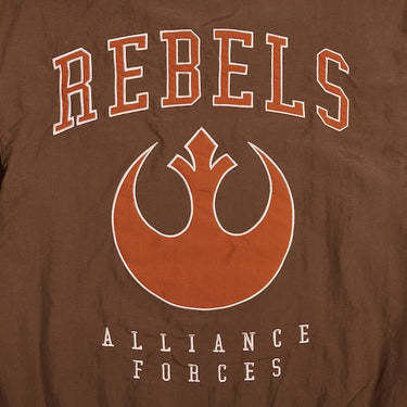 A close up of the back of a Star Wars-themed hooded windbreaker jacket, primarily brown with orange accents. The design features large, bold text reading 'REBELS' across the upper back, with the Rebel Alliance emblem in orange below it. Beneath the emblem, the words 'ALLIANCE FORCES' are printed in a smaller font. The jacket has an elastic waistband and cuffs, with a color-blocked pattern matching the front design.