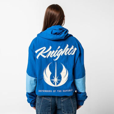A woman standing backwards showing the back of a Star Wars-themed hooded windbreaker jacket, primarily blue with light blue and cream colored accents. The design features large, bold text reading 'Knights' across the upper back, with the Jedi Knight emblem in white with a blue boarder it. Beneath the emblem, the words 'Defenders of the Republic' are printed in a smaller font. The jacket has an elastic waistband and cuffs, with a color-blocked pattern matching the front design.