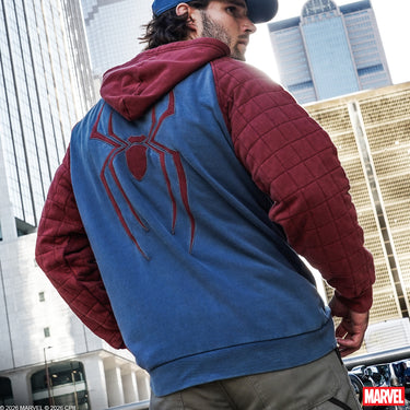 A man wearing a Marvel Spider-Man themed hooded jacket, viewed from behind. The jacket features a blue back panel with a large red Spider-Man web emblem, and quilted burgundy sleeves and hood.