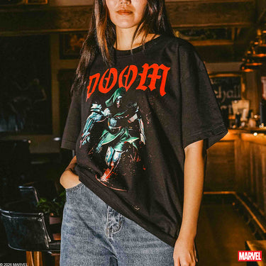 A woman wearing a black graphic t-shirt featuring the word 'DOOM' in red gothic lettering and a green and red comic-style character illustration. The shirt is styled with light-wash blue jeans in a dimly lit, warm-toned indoor setting.