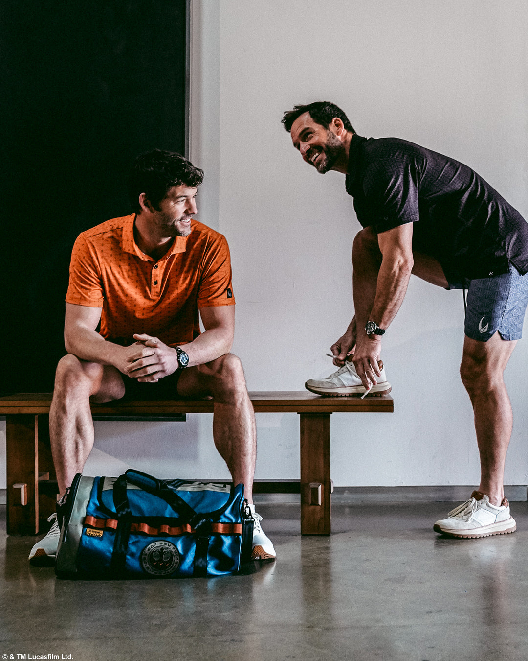 Two men in a casual setting with one sitting on a bench and the other standing, near a blue star wars duffel bag.